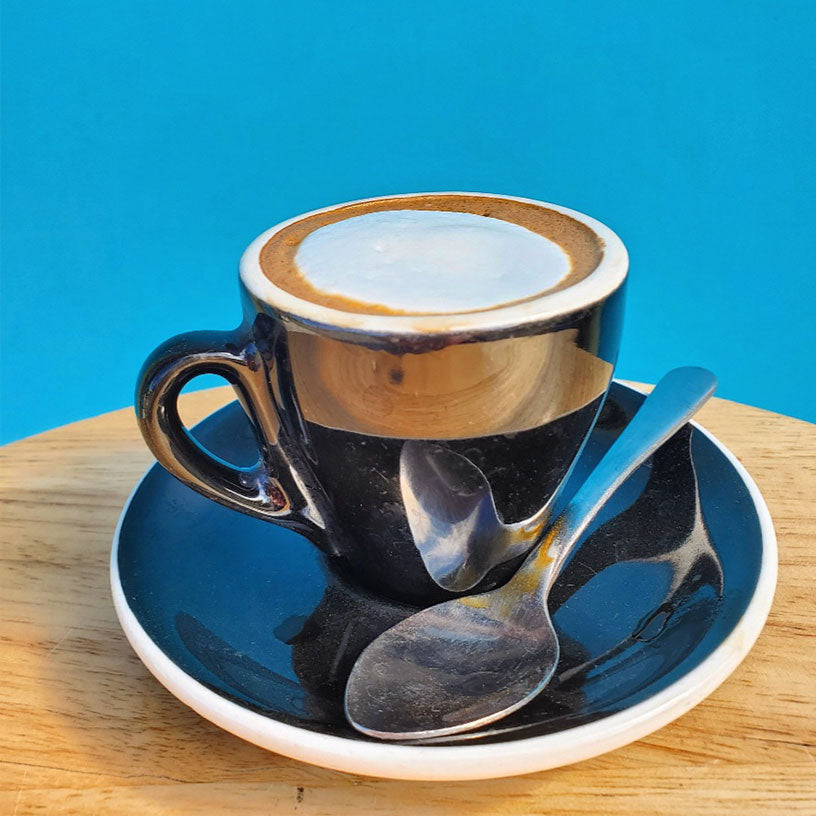 Classic Caffe Ibis machiatto in a small cup with saucer and demitase spoon, against bright blue background and wood table.